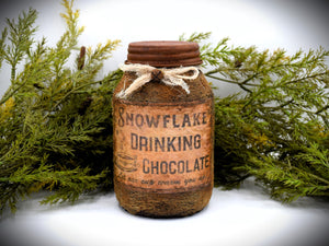 A rustic quart sized pantry jar labeled &quot;Snowflake Drinking Chocolate&quot;, with a vintage-style design and tied with cheesecloth bow, is placed in front of green foliage.