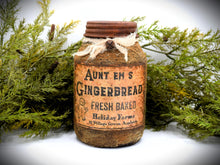 Load image into Gallery viewer, A rustic quart sized pantry jar labeled &quot;Aunt Em’s Gingerbread Fresh Baked&quot; from Holiday Farms, with a vintage-style design and tied with cheesecloth bow, is placed in front of green foliage.