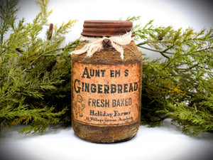 A rustic quart sized pantry jar labeled &quot;Aunt Em’s Gingerbread Fresh Baked&quot; from Holiday Farms, with a vintage-style design and tied with cheesecloth bow, is placed in front of green foliage.