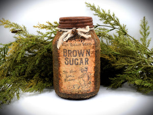 A rustic quart sized pantry jar labeled &quot;Blue Ribbon Brand Brown Sugar&quot;, with a vintage-style design and tied with cheesecloth bow, is placed in front of green foliage.