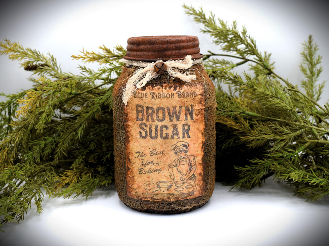 A rustic quart sized pantry jar labeled "Blue Ribbon Brand Brown Sugar", with a vintage-style design and tied with cheesecloth bow, is placed in front of green foliage.