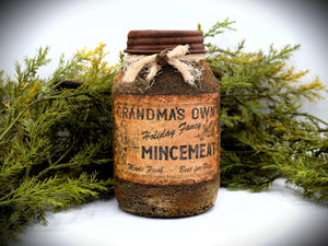 A rustic quart sized pantry jar labeled &quot;Grandma&#39;s Own Holiday Fancy Mincemeat&quot;, with a vintage-style design and tied with cheesecloth bow, is placed in front of green foliage.