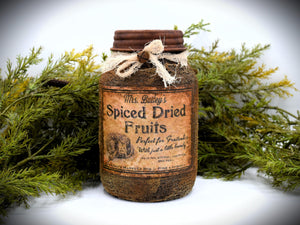 A rustic quart sized pantry jar labeled &quot;Mrs. Bailey&#39;s Spiced Dried Fruits&quot; with a vintage-style design and tied with cheesecloth bow, is placed in front of green foliage.