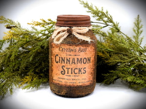 A rustic quart sized pantry jar labeled &quot;Ceylon&#39;s Best Cinnamon Sticks&quot;, with a vintage-style design and tied with cheesecloth bow, is placed in front of green foliage.