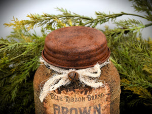 A rustic jar with a rusty lid, is surrounded by greenery. The jar is tied with a piece of tattered cheesecloth, adding a vintage feel to the scene.